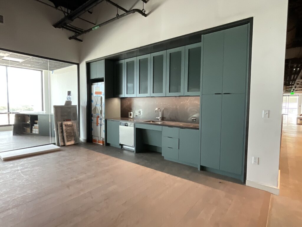 Modern kitchen with teal cabinets and stainless steel appliances, featuring an unfinished open space with natural light from large windows.
